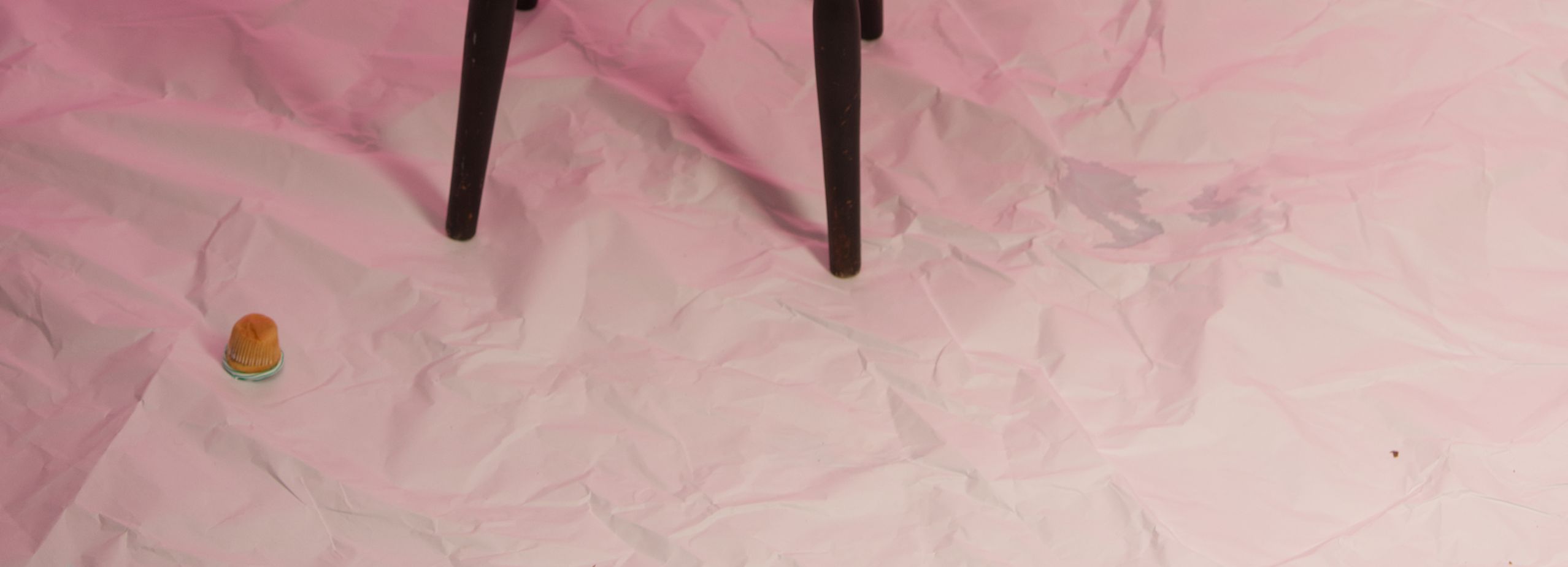 Ein heruntergefallener Muffin auf rosa Stoffboden, daneben Beine eines Barhockers