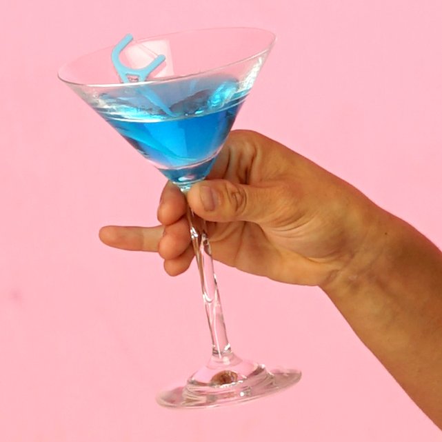 Eine Hand hält ein Martiniglas gefüllt mit einer blauen Flüssigkeit. Im Glas schwimmt ein Zahnseidestäbchen.